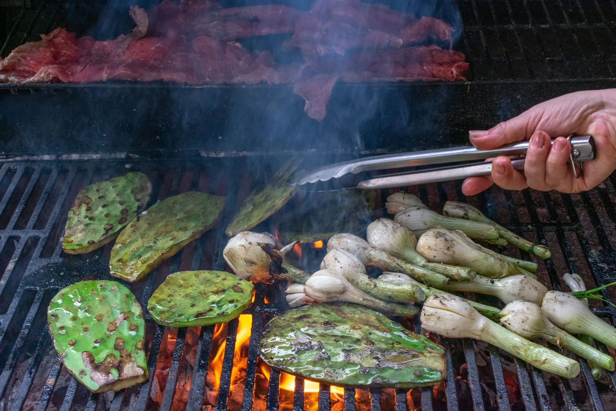 Native foods to Baja California Sur. Closeup of carne asada, nopales and onions being grilled - one hand of the cook showing
